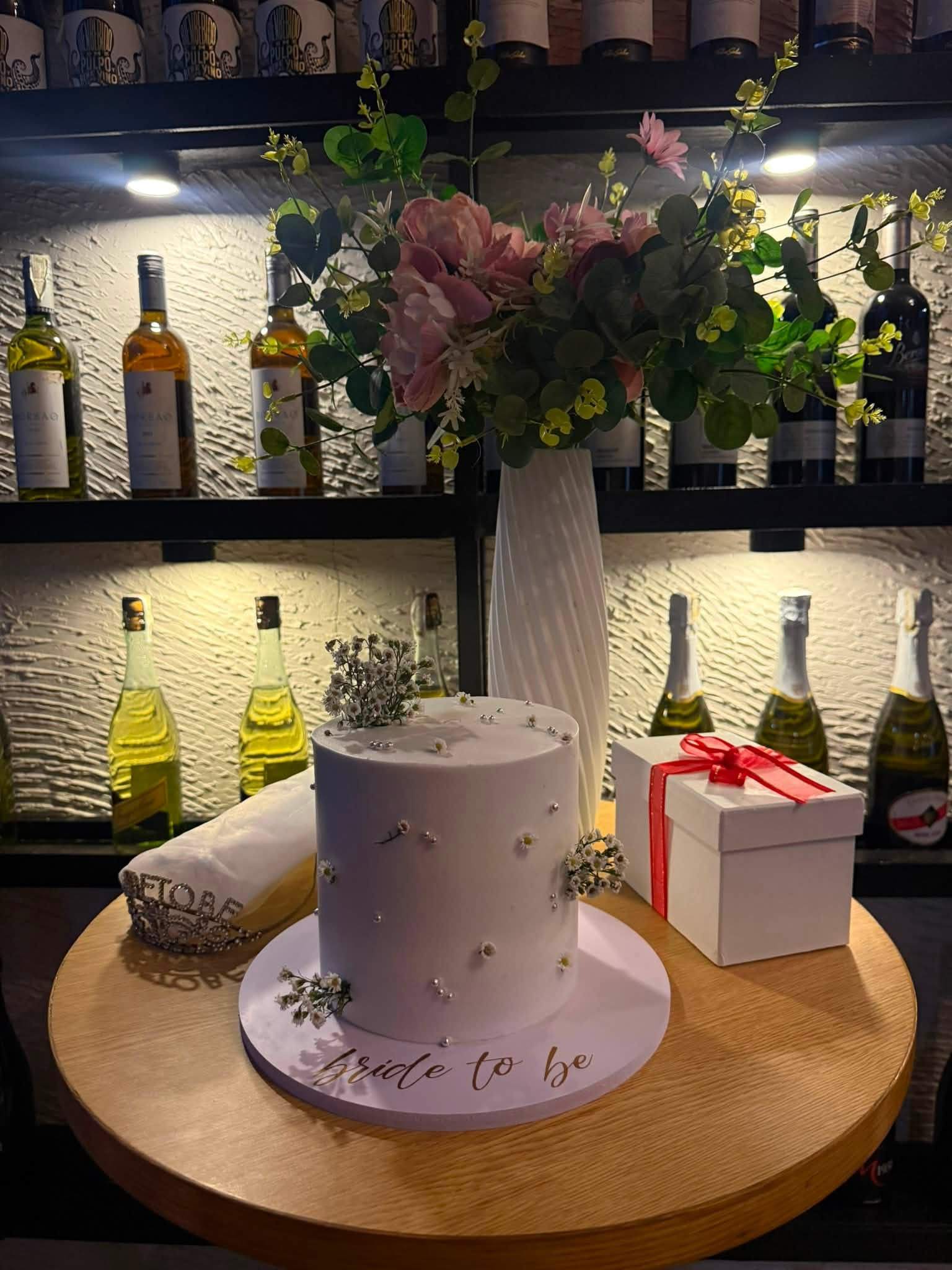 A white celebration cake on a wooden stand beside gift boxes and flowers