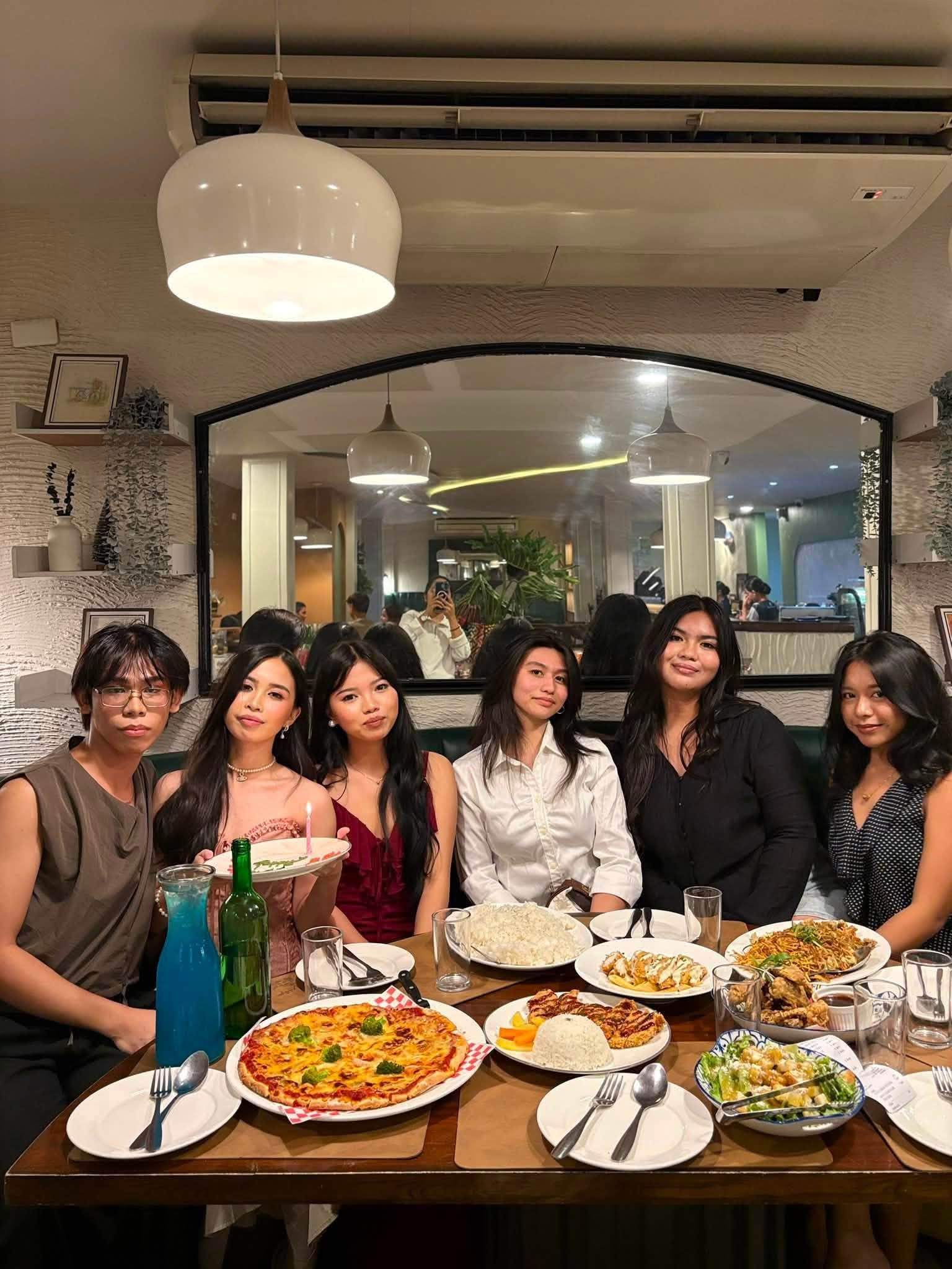 A group of friends smiling around a table of pizza, pasta and drinks