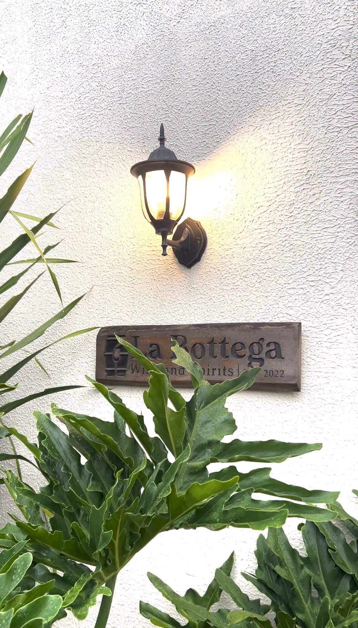 A small wooden La Bottega sign and lantern on a white wall framed by leaves