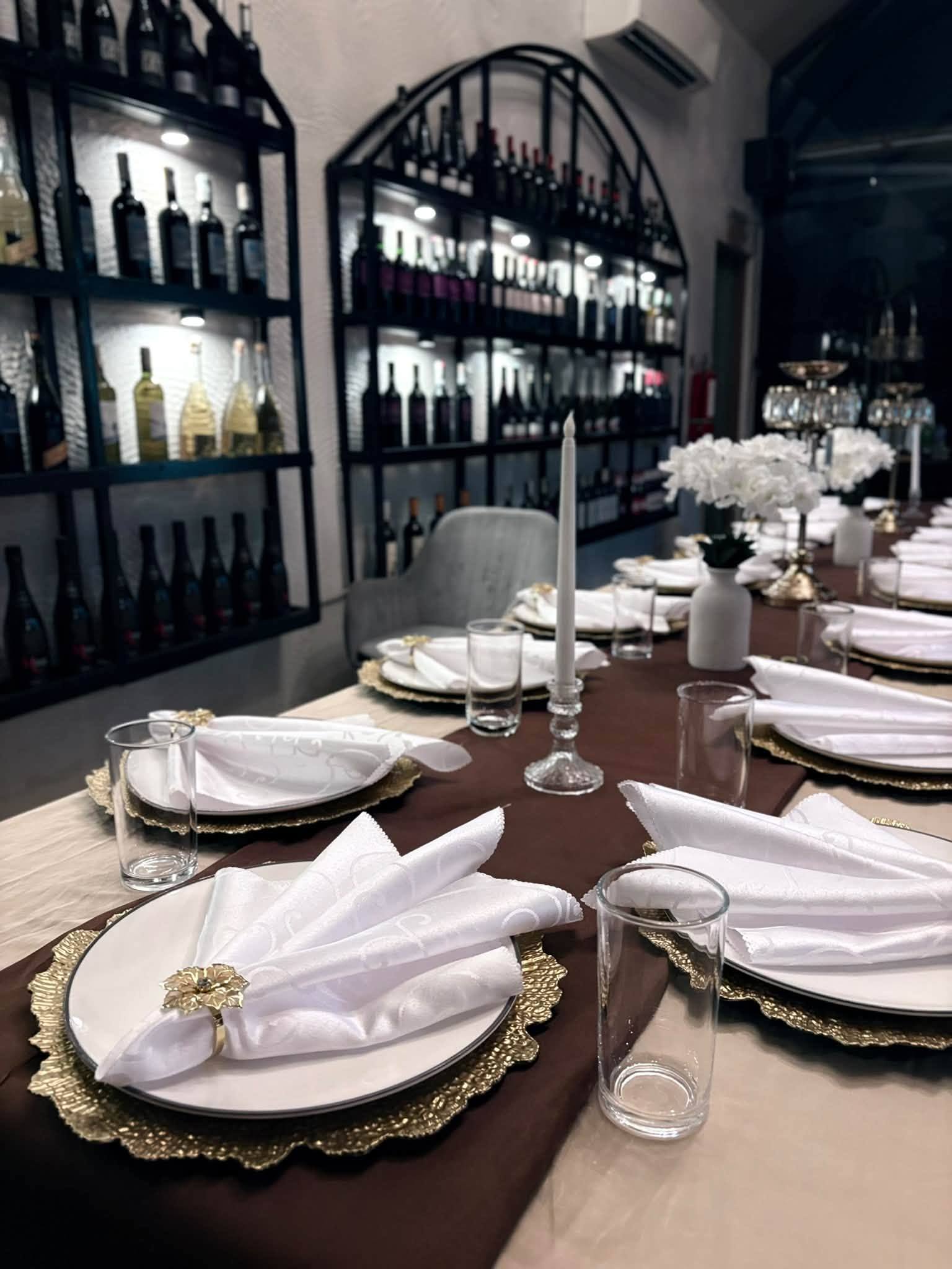 A table laid with white napkins and gold chargers in front of illuminated wine shelves