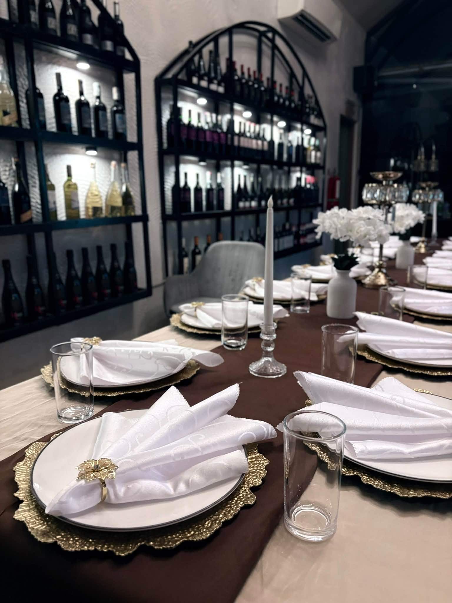A table laid with white napkins and gold chargers in front of illuminated wine shelves