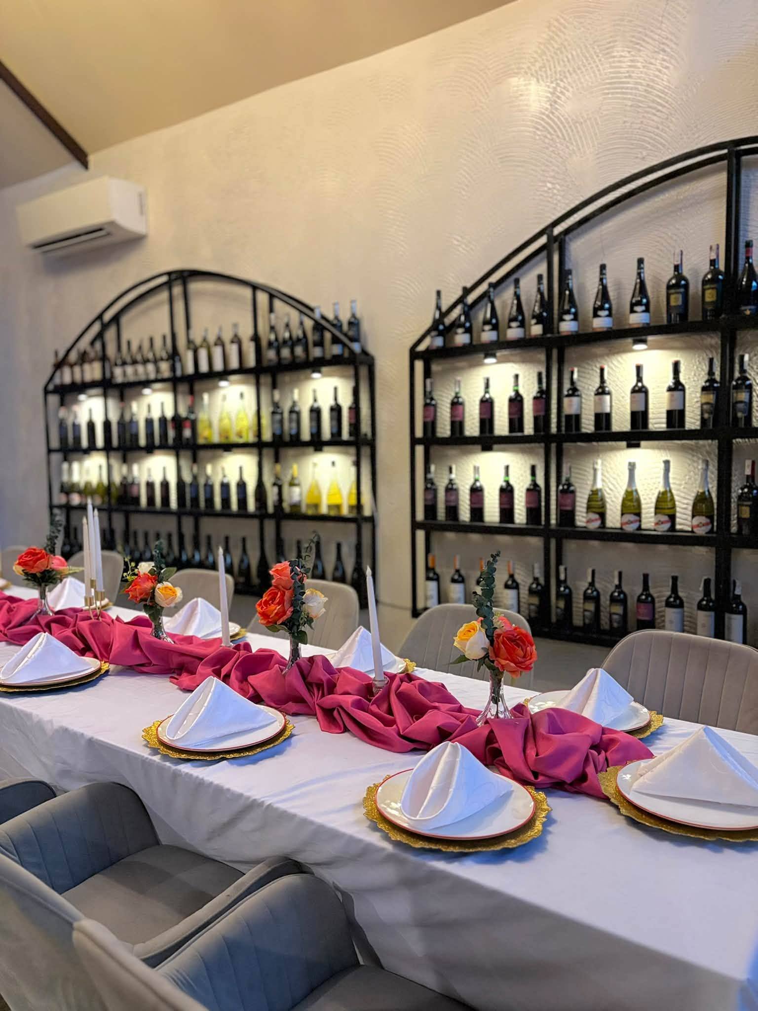 Arched black shelves of wine bottles lit from behind above a long table set with pink linens and flowers