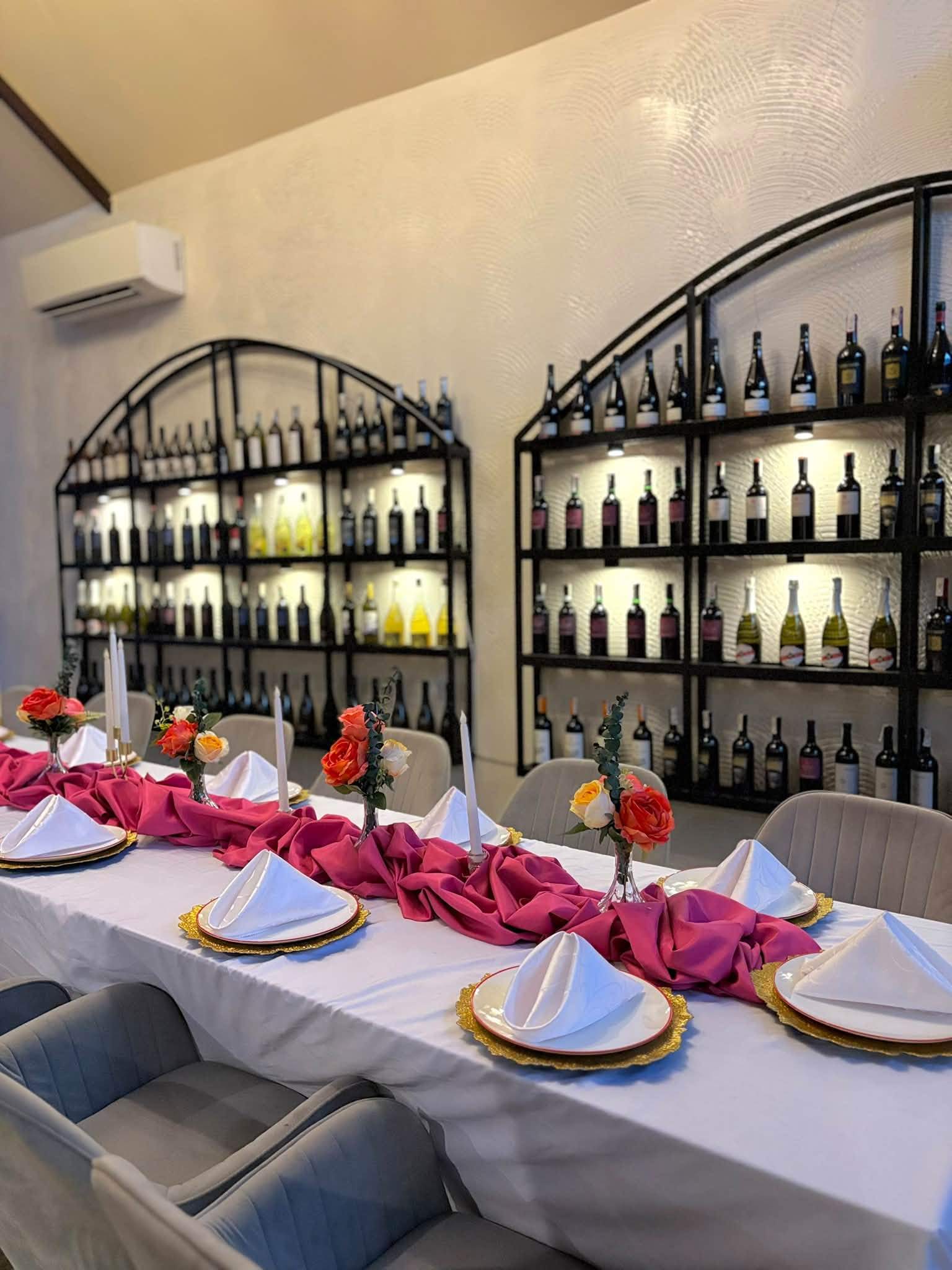 Arched black shelves of wine bottles lit from behind above a long table set with pink linens and flowers