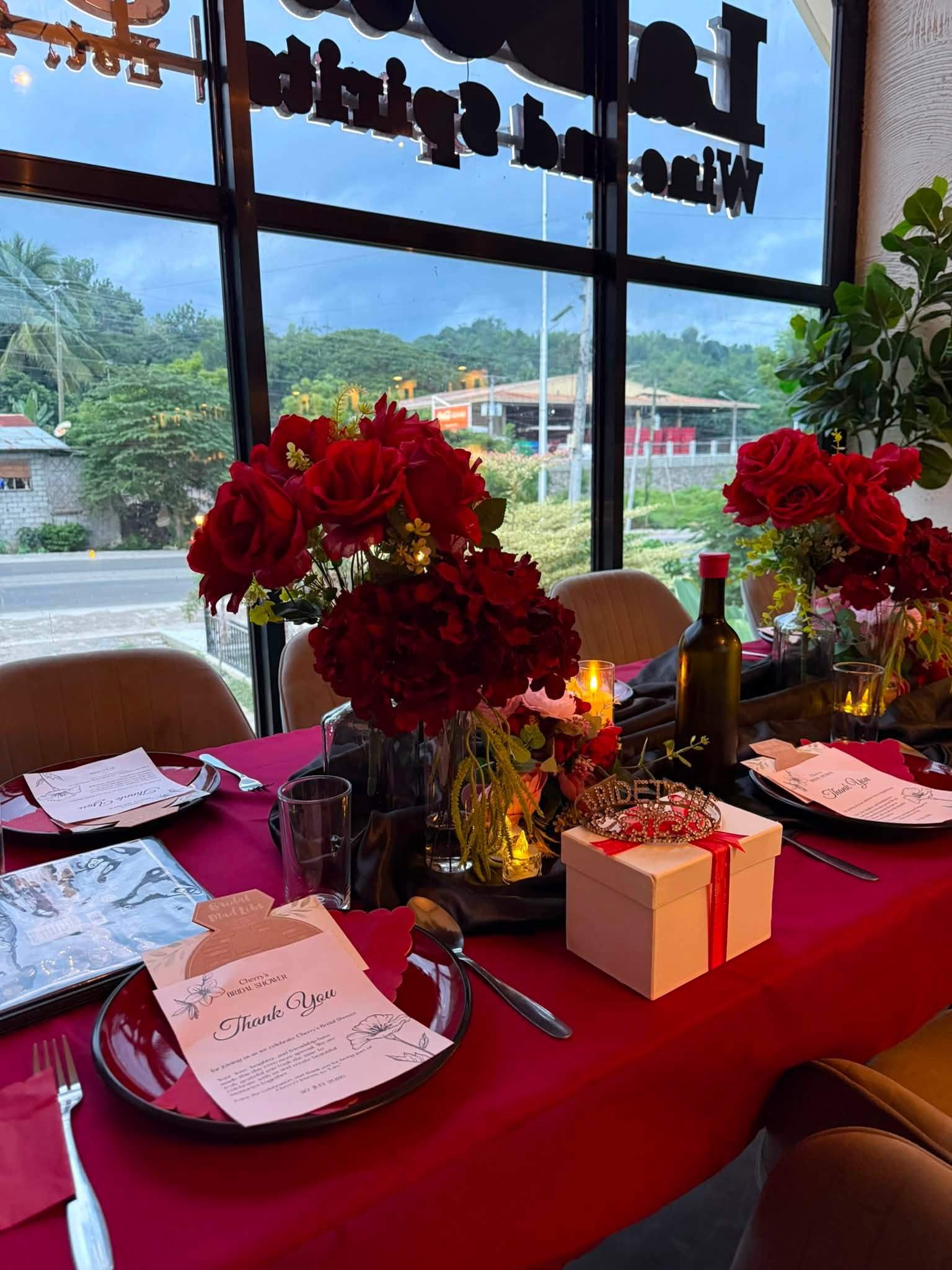 Red roses and a table set for two by the window, with greenery outside